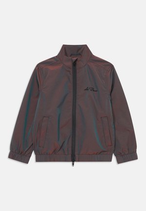 Short jacket in dark metallic fabric with high collar, zip closure, and elastic cuffs. Features side pockets and a subtle logo on the chest.