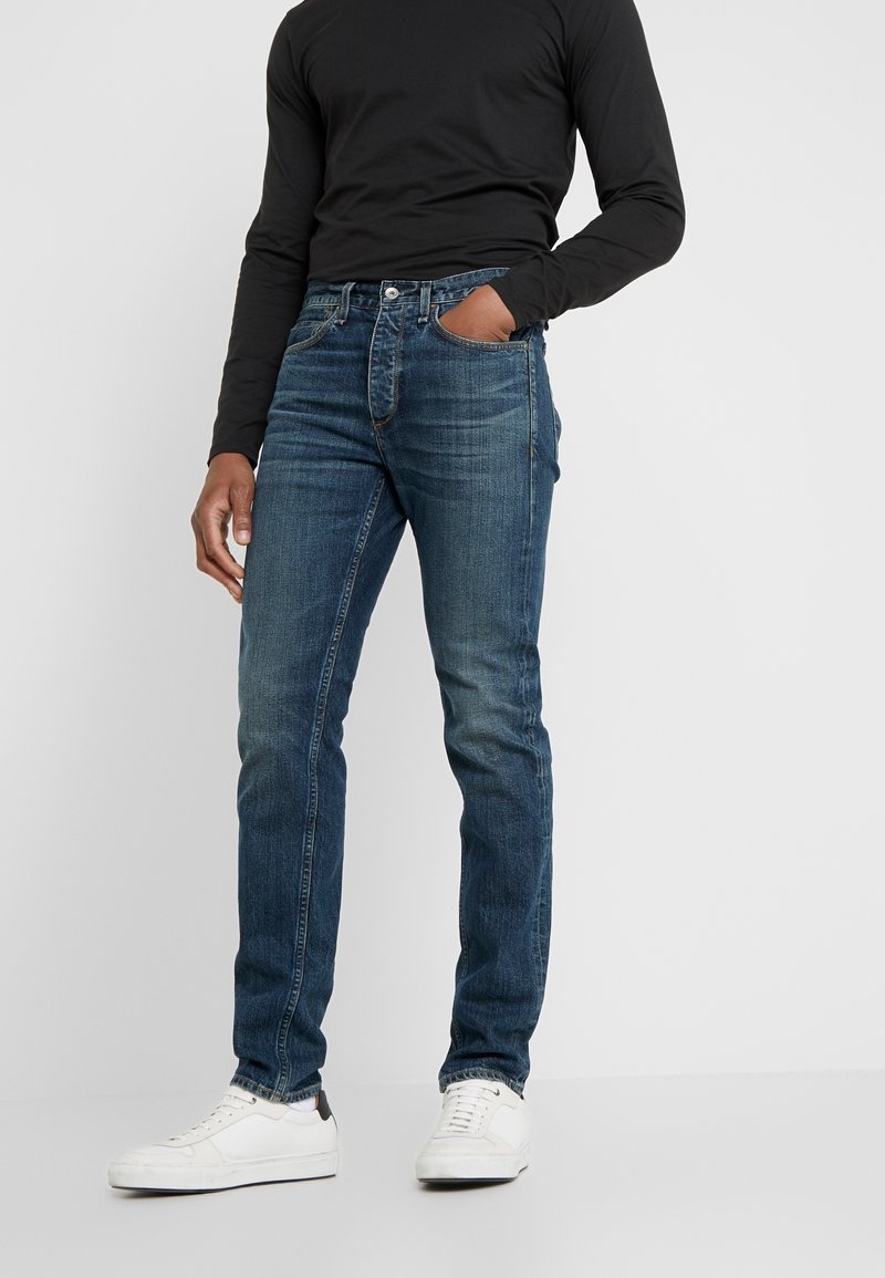 Jeans de mezclilla azul oscuro con corte ajustado, que presentan un diseño clásico de cinco bolsillos, un ligero desgaste y un cierre estándar de botón y cremallera.