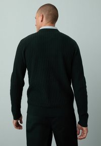 Dunkelgrüner Strickpullover mit strukturiertem Rippenmuster, langen Ärmeln und Rundhalsausschnitt, kombiniert mit schwarzen Hosen. Ansicht von hinten.