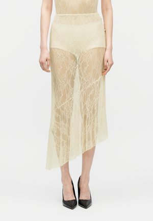 Γυναίκα που φοράει μπεζ διαφανές δαντελένιο φόρεμα με ασύμμετρο τελείωμα και μαύρες γόβες με μυτερές μύτες, στέκεται μπροστά σε λευκό φόντο.