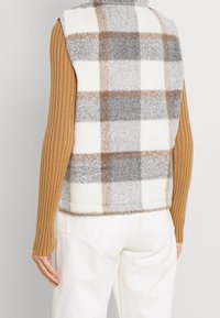 Westen im karierter Design mit grauen, weißen und braunen Farben, aus Wollmaterial und mit hohem Kragen. Geripptes Langarmoberteil in warmem Braun.