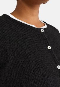 Cardigan noir texturé avec un col rond orné d'une bordure blanche et de deux boutons blancs pour la fermeture. La vue en gros plan met en évidence les détails du tissu.
