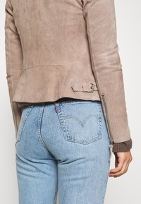 Svetle béžová semišová bunda s perforovaným vzorem, páskem v pase a detaily na zip, nošená s světlými modrými džínami s logem na zadní kapse.