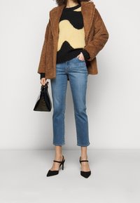 Chaqueta de pelaje sintético marrón, suéter con estampado negro y amarillo, jeans cortos de mezclilla azul claro, zapatos de tacón negros y un pequeño bolso negro.