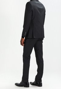 Completo nero su misura con giacca strutturata e pantaloni slim-fit. Caratterizzato da una superficie liscia, cuciture sottili e scarpe stringate nere.