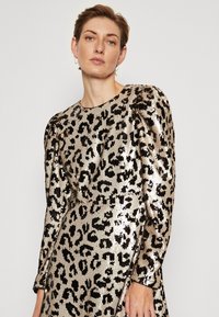 Robe en imprimé léopard avec des sequins noirs et argentés, manches longues bouffantes, col rond et coupe ajustée. Texture scintillante et motif audacieux.