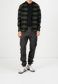 Camicia di flanella a quadri verdi e neri sopra una maglietta nera, abbinata a pantaloni cargo scuri e sneakers bianche con dettagli grigi.