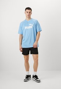Lyseblå t-shirt med korte ærmer og hvid "PUMA" logo på fronten, kombineret med sorte shorts. Modellen bærer sorte og hvide sneakers med hvide sokker.