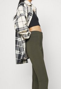 Rutig flanellskjorta i kräm och marinblå, lagd över en svart crop top, parat med olivgröna figurnära leggings som visar en slät textur.