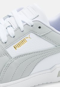 Gros plan sur une chaussure Puma blanche et gris clair avec des lacets blancs, un logo Puma doré et des détails en cuir texturé.