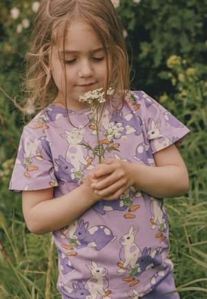 Jauna meitene ar gaiši brūniem matiem, nēsājot violets krāsas krekliņu ar zaķu attēlu, maigi tur un smaržo baltas savvaļas puķes ārā.
