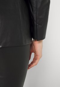 Černá kožená bunda s hladkou texturou, s přizpůsobeným střihem a viditelnými švy. Ruka spočívající na odpovídající černé sukni.