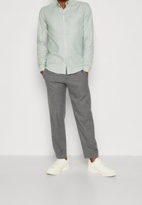 Jasnozielona koszula na długi rękaw zapinana na guziki, szare zwężane spodnie i białe sneakersy, stojące na jednolitym tle. Gładka tkanina, casualowy krój.