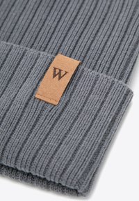 Cappello a maglia grigio a coste con risvolto pieghevole e una targhetta in pelle marrone chiaro con la lettera "W" in rilievo. Texture morbida, design casual.