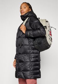 Žena s pletenicom nosi crnu puf jaknu i drži ruksak na ramenu, samouvjereno se smiješeći na svijetloj pozadini.