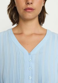 Blusa azzurro chiaro con pieghe verticali, scollo a V e sei bottoni blu davanti. Tessuto morbido e liscio con una texture delicata.