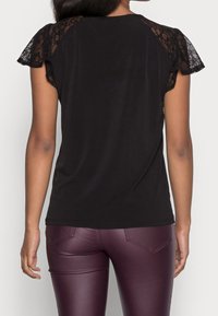 A woman’s back is facing the camera, wearing a black top with lace sleeves and maroon fitted pants, against a plain background.