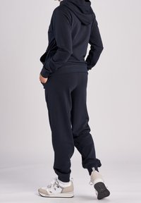 Mörkblå hoodie med foder i huvan och fickor, tillsammans med matchande joggers. Modellen bär vita och beiga sneakers. Slät textur.