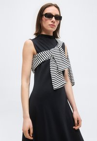 Femme portant une robe noire sans manches, des lunettes de soleil rectangulaires noires, et un pull rayé noir et blanc posé sur les épaules et noué devant.