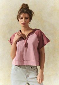Korte mouwen, cropped top in roze met een donkerdere roze schouderpas. Heeft een knopenlijst en rafelige randen. Gecombineerd met lichte blauwe jeans.