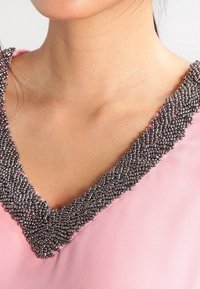 Abbigliamento rosa con scollatura a V adornato con perline metalliche. Texture liscia con decorazioni di perline lungo il collo, evidenziando i dettagli.