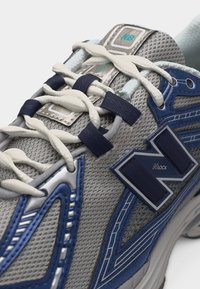 Κοντινό πλάνο γκρι και μπλε αθλητικού παπουτσιού New Balance με λευκά κορδόνια περασμένα από ναυτικά οπές και λογότυπο "Nlock" στο πλάι.