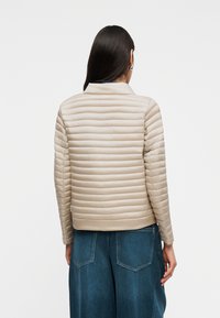 Γυναίκα με μακριά σκούρα μαλλιά που φοράει μπεζ μπουφάν με ραφές και μπλε τζιν παντελόνι, γυρισμένη πλάτη σε απλό λευκό φόντο.