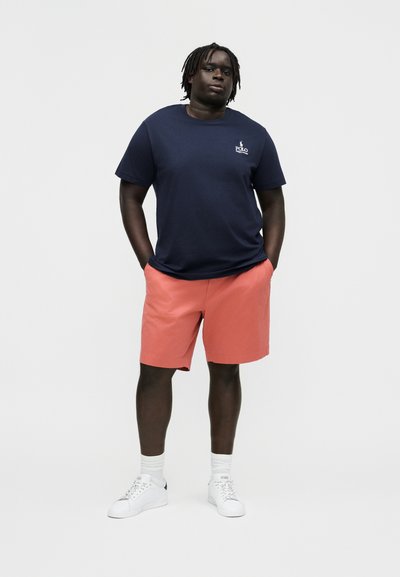 Mann steht mit Händen in den Taschen, trägt ein marineblaues Poloshirt, korallenrote Shorts, weiße Socken und weiße Turnschuhe vor einfachem Hintergrund.