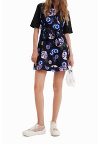 Czarna sukienka z kolorowymi okrągłymi wzorami, krótkie rękawy, talia z paskiem oraz spódnica w warstwach; zestawiona z białymi sneakersami i białą torebką.