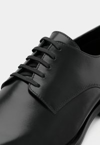 Chaussure en cuir noir à finition lisse, dotée d'œillets et de lacets noirs. Le design comprend des accents de couture subtils le long de la tige.