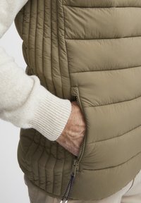Olivengrüner aufgepusterter Weste mit gestepptem Muster, seitlichem Reißverschlussfach und hohem Kragen. Strickbeige Pulloverwärmer sichtbar.