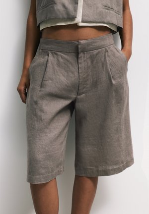 Personne portant un short en lin gris taille haute, ample, avec des plis devant et des poches latérales, assorti d'un gilet court coordonné.