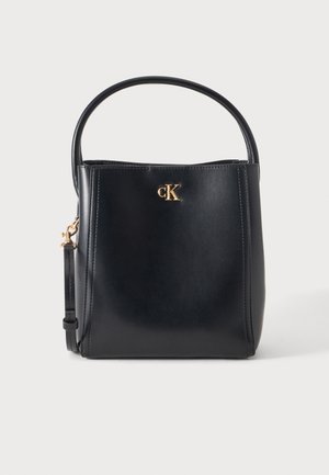 Schwarze strukturierte Handtasche mit rundem Henkel, goldenem CK-Logo auf der Vorderseite und goldener Schnalle an der Seite, vor weißem Hintergrund.