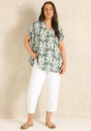 Kvinde iført grøn mønstret løstsiddende top, hvide korte bukser og beige sandaler, stående op ad en ensfarvet beige væg.