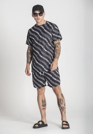 Camicetta nera a maniche corte e pantaloni corti abbinati con testo del logo bianco ripetuto. Il tessuto ha una texture liscia, completata da semplici sandali neri.