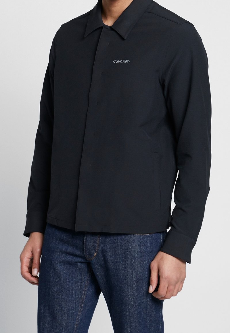 Sort kravejakke med knap- frontdesign, lavet af glat stof. Har et lille hvidt "Calvin Klein" logo på brystet.