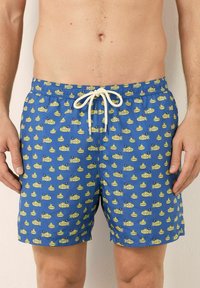 Des maillots de bain bleus avec un motif de sous-marin ludique en jaune, dotés d'un cordon blanc et d'une taille élastique. Tissu léger.