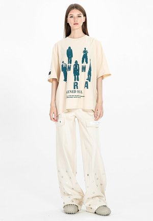 Joven mujer está de pie con una camiseta gráfica beige de gran tamaño, pantalones cargo de pierna ancha con puntos plateados y zapatos gruesos texturizados sobre fondo blanco.