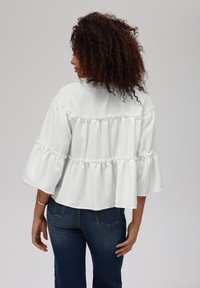 Hvit, teksturert topp med volanger og vide ermer, kombinert med mørkeblå jeans. Bakfra viser den en firkantet silhuett.