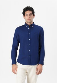 Camisa de botones azul oscuro con mangas largas, hecha de tela suave. Presenta un cuello con solapa y un pequeño logo en el pecho.
