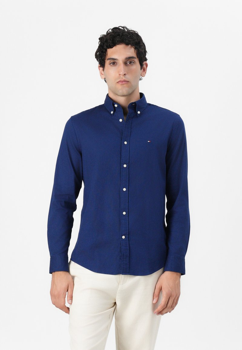 Camisa de botones azul oscuro con mangas largas, hecha de tela suave. Presenta un cuello con solapa y un pequeño logo en el pecho.