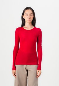 Red long-sleeve top made of lightweight fabric with a round neckline, fitted design, and smooth texture. Paired with beige loose-fitting trousers.