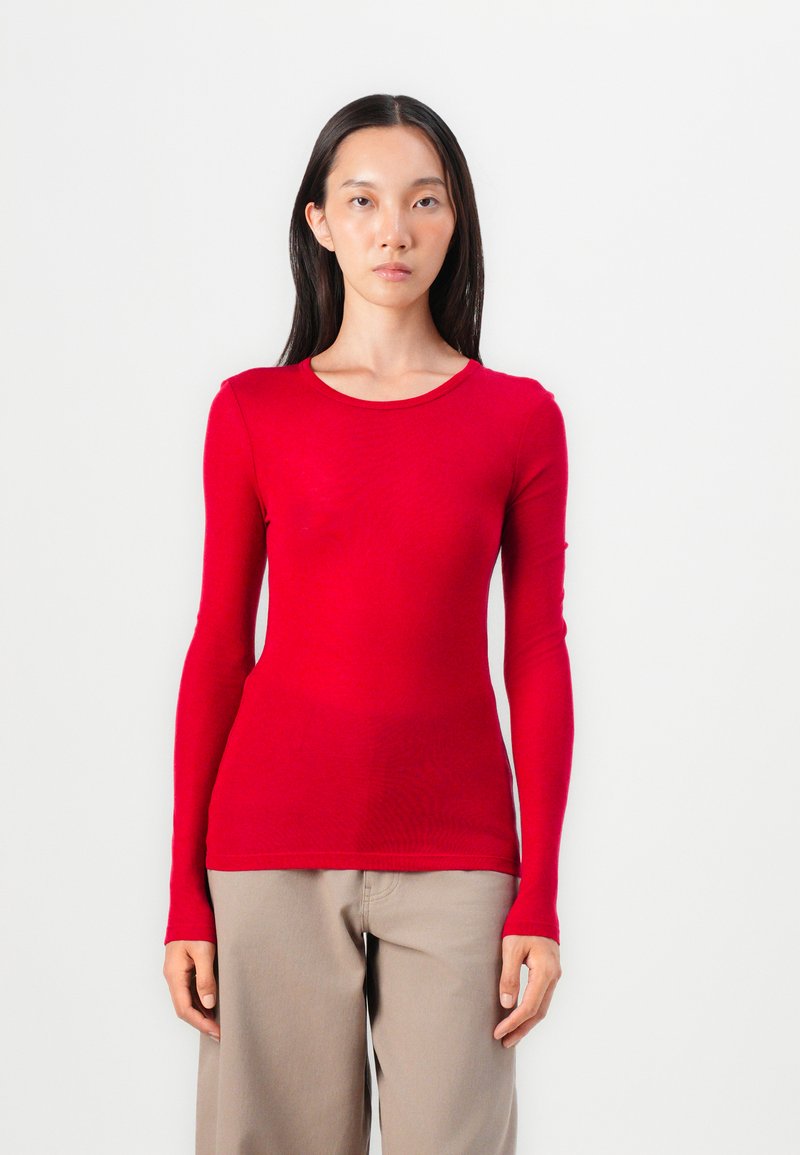 Red long-sleeve top made of lightweight fabric with a round neckline, fitted design, and smooth texture. Paired with beige loose-fitting trousers.