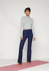 Vrouw die blootsvoets op een roze yogamat staat, gekleed in een lichtgrijze lange mouwen top en marineblauwe aansluitende broek tegen een effen achtergrond.