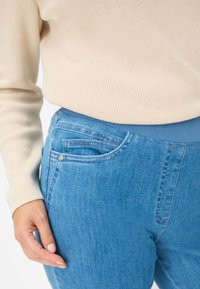 Jean bleu clair avec une texture en denim lisse, comportant une poche avant et une taille élastique, associé à un pull côtelé de couleur crème.