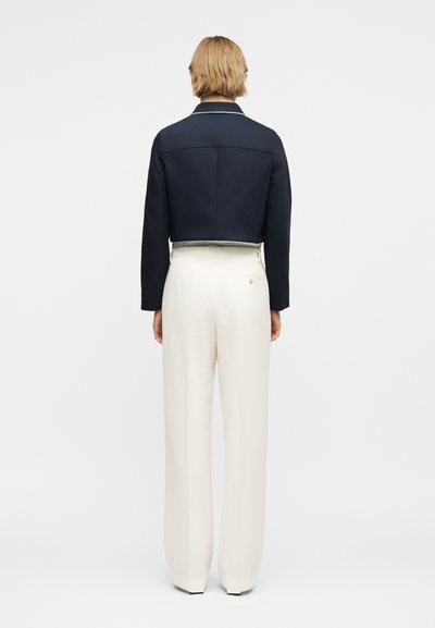 Άτομο με κοντά ξανθά μαλλιά που φοράει ναυτικό κοντό μπουφάν και λευκά, ψηλόμεσα, φαρδιά παντελόνια, στέκεται κοιτώντας προς ένα λευκό φόντο.