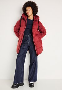 Rød pufferjakke med hætte, med lynlås foran, parret med brede mørke denimjeans og sorte chunky støvler.