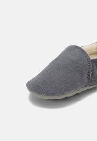 Chausson en tissu gris doux avec bout fermé et doublure en peluche claire, vu de profil sur fond blanc.