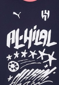 Al-Hilal logo, Puma logo, stiliseret tekst "AL-HILAL," stjerner og en fodboldgrafik på mørk stofbaggrund.