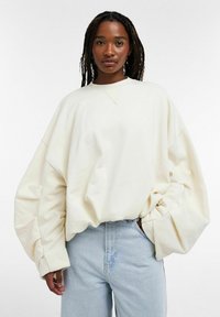 Femme aux longs cheveux tressés portant un sweat-shirt blanc oversize et un jean bleu clair, debout devant un fond blanc uni.